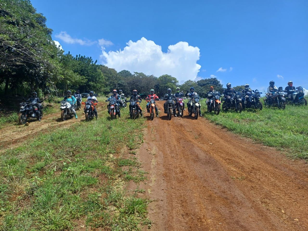 Unos quince motociclistas en fila sobre un camino de tierra roja, bajo cielo azul.