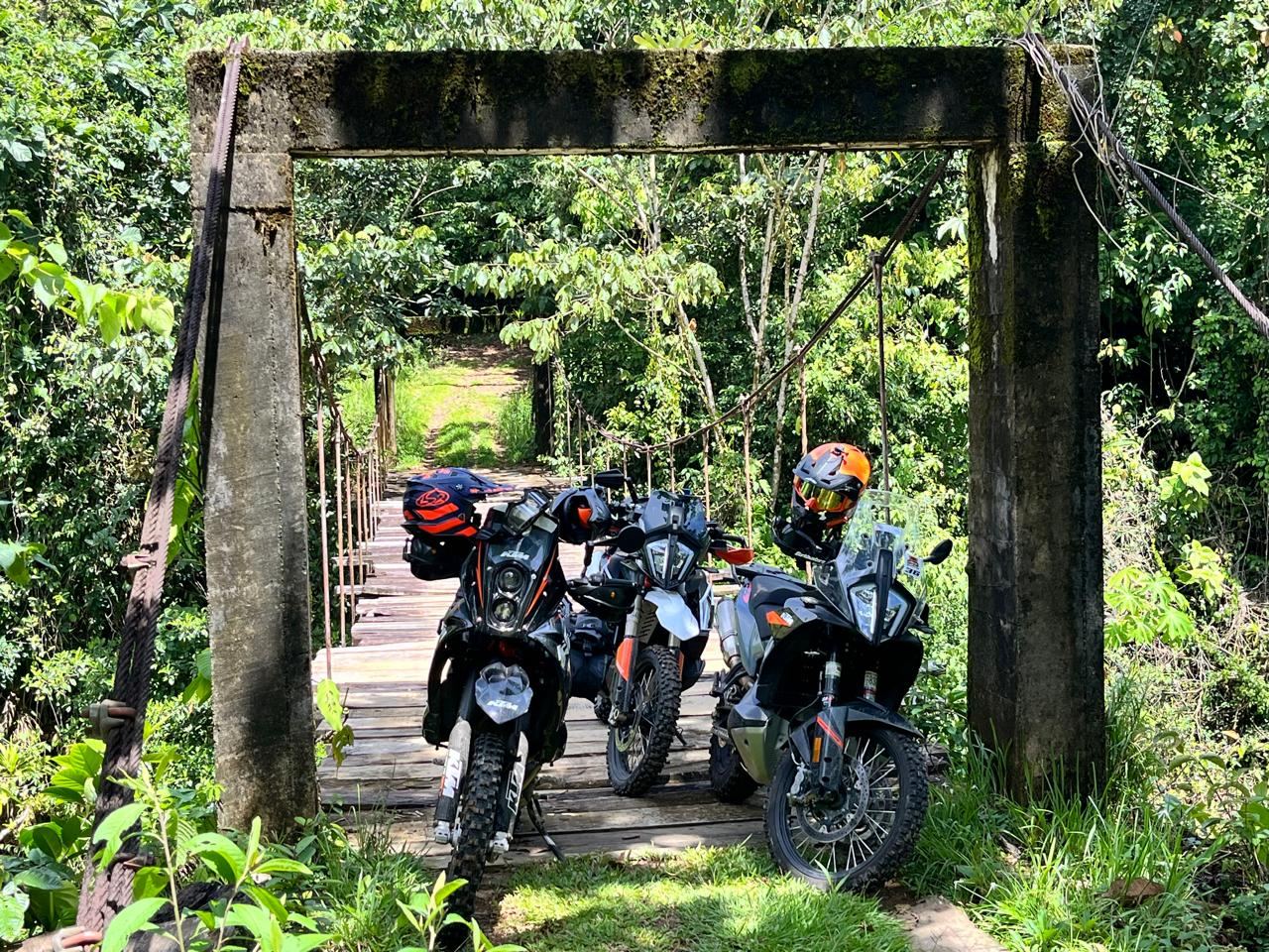 Tres motos KTM con los cascos puestos en un puente colgante de madera, en el bosque.
